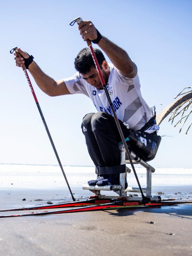 In preparation for the Milano Cortina 2026 Paralympic Winter Games, a male Para athletes trains in a sit-ski on the beach, propelling forward using two poles.